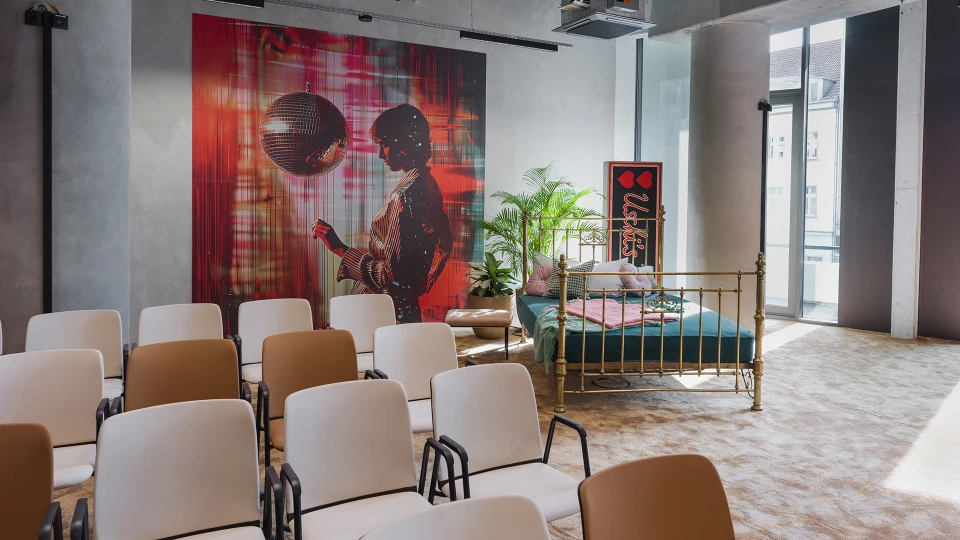 Reihe von weißen und braunen Stühlen in einem Seminarraum mit Teppichboden, im Hintergrund eine Wand mit einem großen roten Kunstmotiv und einem goldenen Bettgestell.