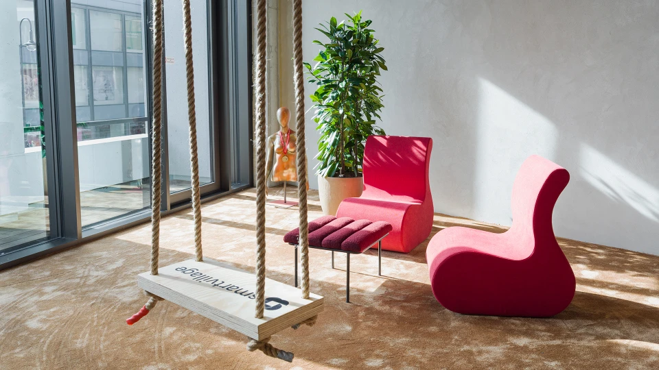 Ecke eines Raumes mit zwei pinken Sesseln, einem Hocker, einer Schaukel mit 'smartvillage'-Logo und einer großen Pflanze, Tageslicht durch bodentiefe Fenster.