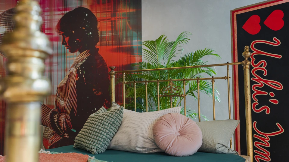 Detailansicht eines goldfarbenen Bettgestells mit Kissen, im Hintergrund das rote Kunstmotiv und grüne Pflanzen.