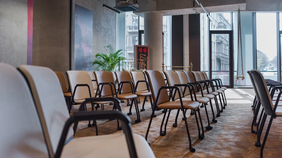 Perspektive durch mehrere Reihen weißer und brauner Stühle in einem Seminarraum, im Hintergrund das Kunstmotiv und das goldfarbene Bett.