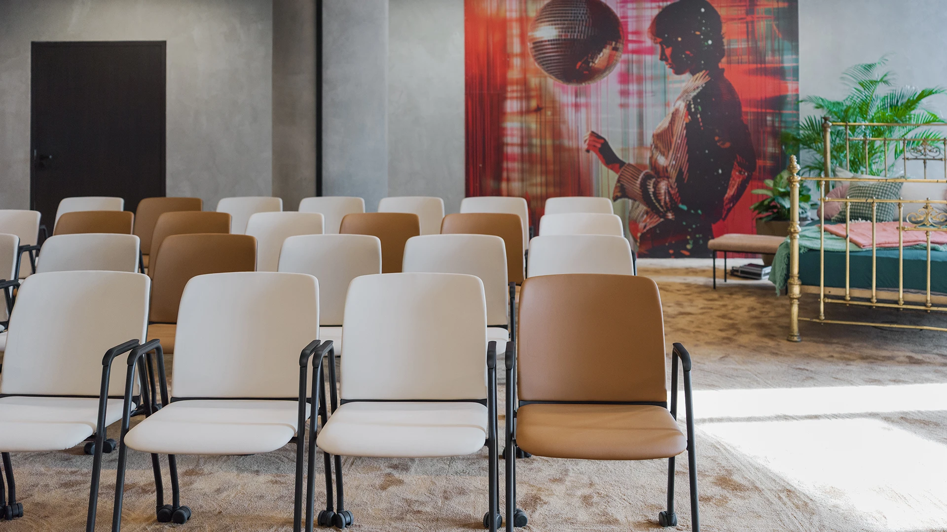 Seminarraum mit Fokus auf die ersten Stuhlreihen, weiß und braun, Hintergrund zeigt eine Wand mit Kunstmotiv und ein goldenes Bettgestell.