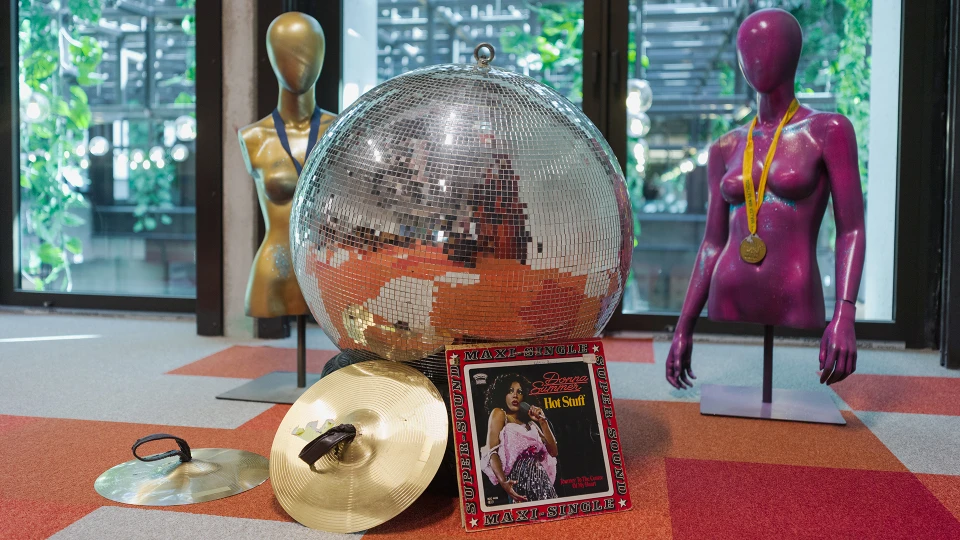 Große silberne Diskokugel hängt von der Decke über dem Konferenzbereich des Disco Stage, Stuhlreihen und bunte Teppichfliesen sichtbar.