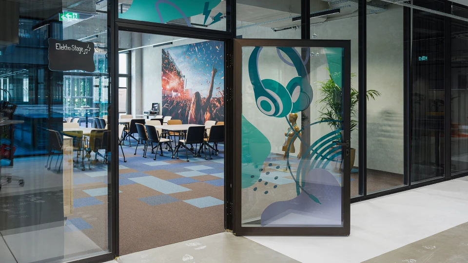 Eingang zur Elektro Stage mit Glasfront und geöffneter Glastür mit aufgedruckten türkisfarbenen Kopfhörern, Blick ins Innere mit Stühlen und bunter Wandgestaltung.