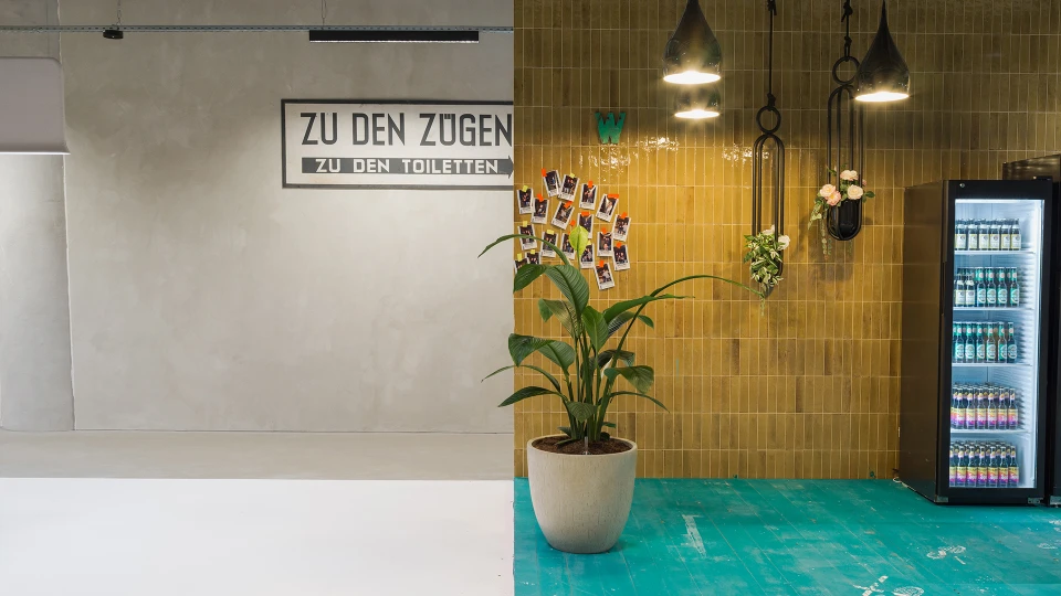 Wandgestaltung mit Vintage-Bahnhofsschild „ZU DEN ZÜGEN / ZU DEN TOILETTEN“, daneben Kühlschrank, Pflanzen und Polaroid-Fotowand.