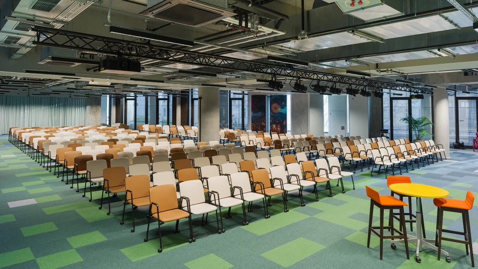Conference room with tiered row seating in light colors; in the foreground a yellow standing table with two orange stools.