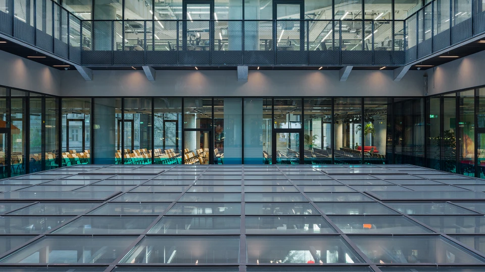View through a courtyard with a glass roof to an event area, where rows of chairs are visible through floor-to-ceiling windows.