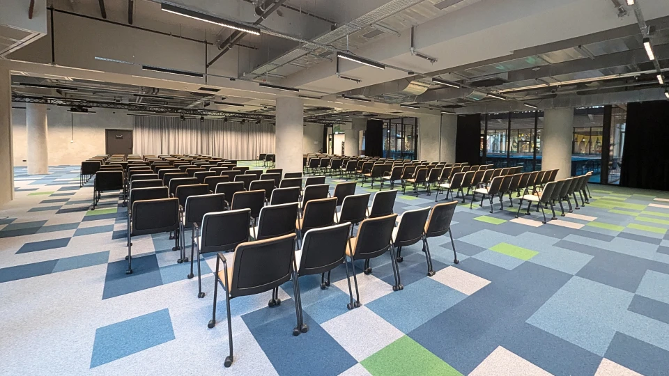 Großer, moderner Konferenzraum mit vielen Reihen schwarzer Stühle, gerichtet auf eine Bühne. Der Raum hat eine bunte Teppichboden und Beleuchtung von der Decke.