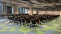Empty conference room with rows of black chairs on a green-blue carpet, ceiling lighting, and an abstract painting on the wall.