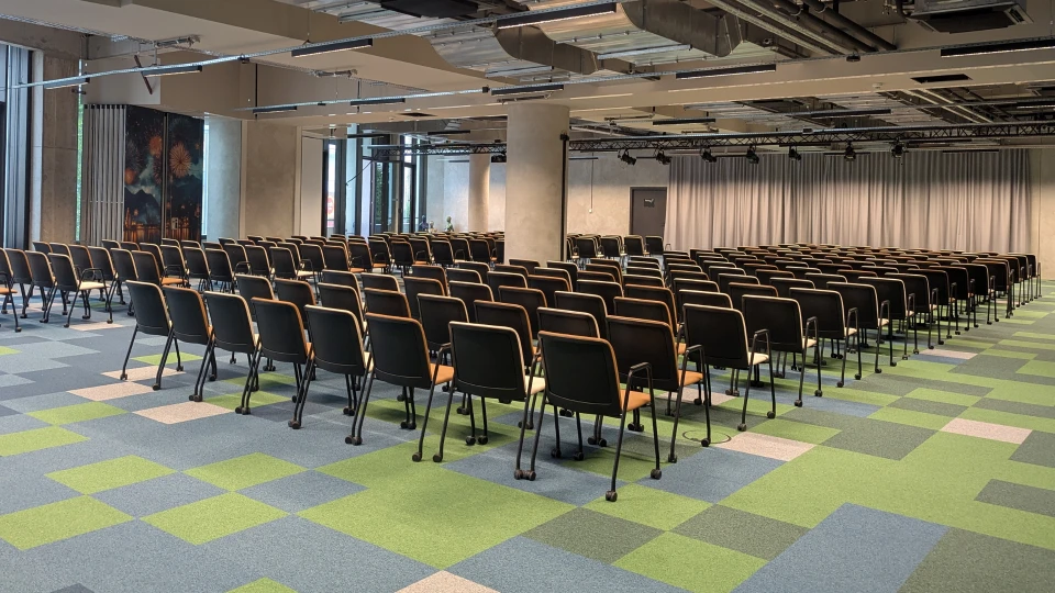 Empty conference room with rows of black chairs on a green-blue carpet, ceiling lighting, and an abstract painting on the wall.