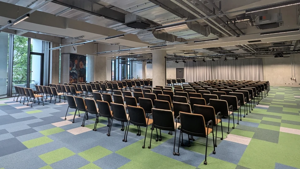 Modern, spacious conference hall with rows of black chairs, green-blue checkered carpet, and exposed ceiling installations