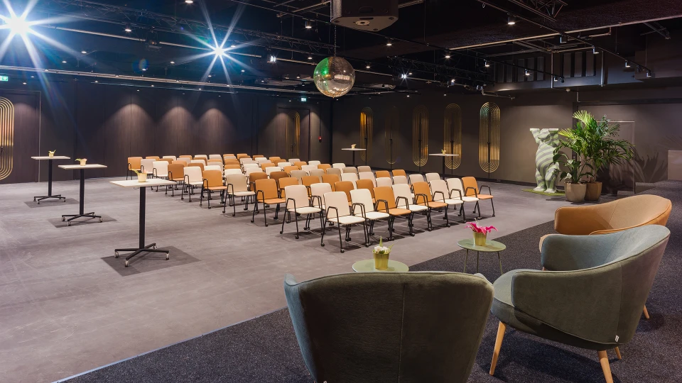 Modern gestalteter Konferenzraum mit Reihen von beige-braunen Stühlen, Stehtischen, Grünpflanzen und einer Diskokugel an der Decke