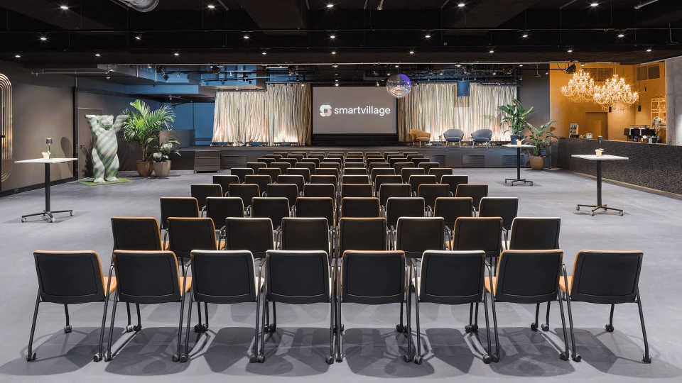 Konferenzraum mit Reihen von schwarzen Stühlen, gerichtet auf eine Bühne mit dem Schriftzug 'smartVillage', dekorativer Beleuchtung und Pflanzen