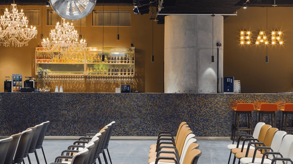 Stilvoller Innenraum eines Bars mit Kronleuchtern, blauer Mosaiktheke, Barhockern und einer beleuchteten 'BAR' Schriftzug