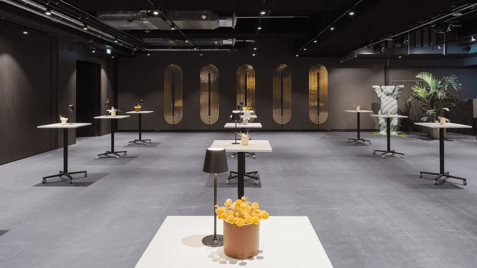 Moderne Ausstellungshalle mit mehreren Tischen, auf denen künstlerische Objekte präsentiert werden, beleuchtet durch stilvolle Wandlampen im Hintergrund.