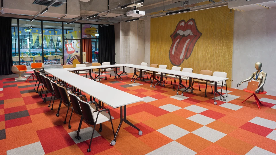 Meetingraum mit U-förmig gestellten Tischen, orangerotem Teppich, markanter Zungen-Grafik an der Wand und Sitzfigur mit Schallplatte.