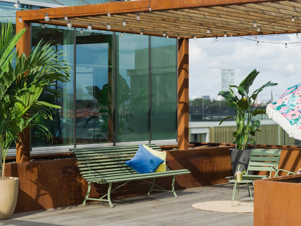 Cozy terrace of a modern building with green metal benches, large potted plants, and a colorful umbrella.