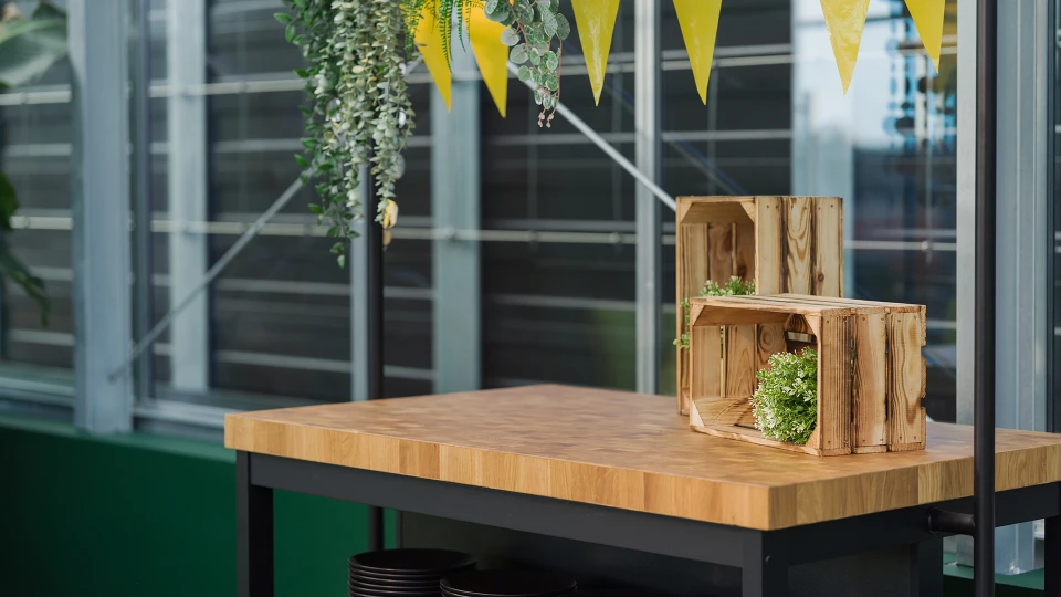 Holztisch mit dekorativen Holzkisten und grünen Pflanzen auf einer Terrasse, geschmückt mit gelben Wimpeln und moderner Glasfassade im Hintergrund.