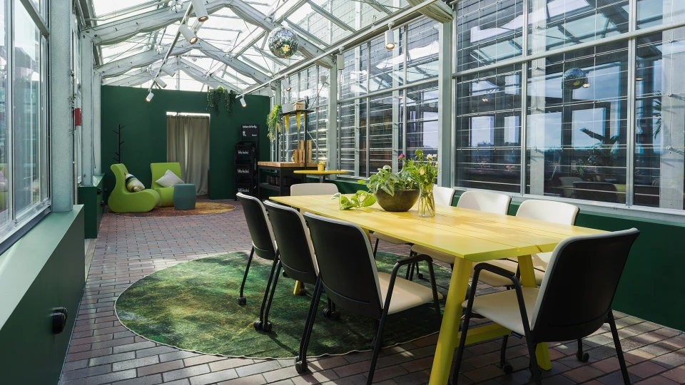 Ein helles Wintergarten-Interieur mit einem langen gelben Tisch und schwarzen Stühlen, einer grünen Sitzbereich, Topfpflanzen und Glasdach.