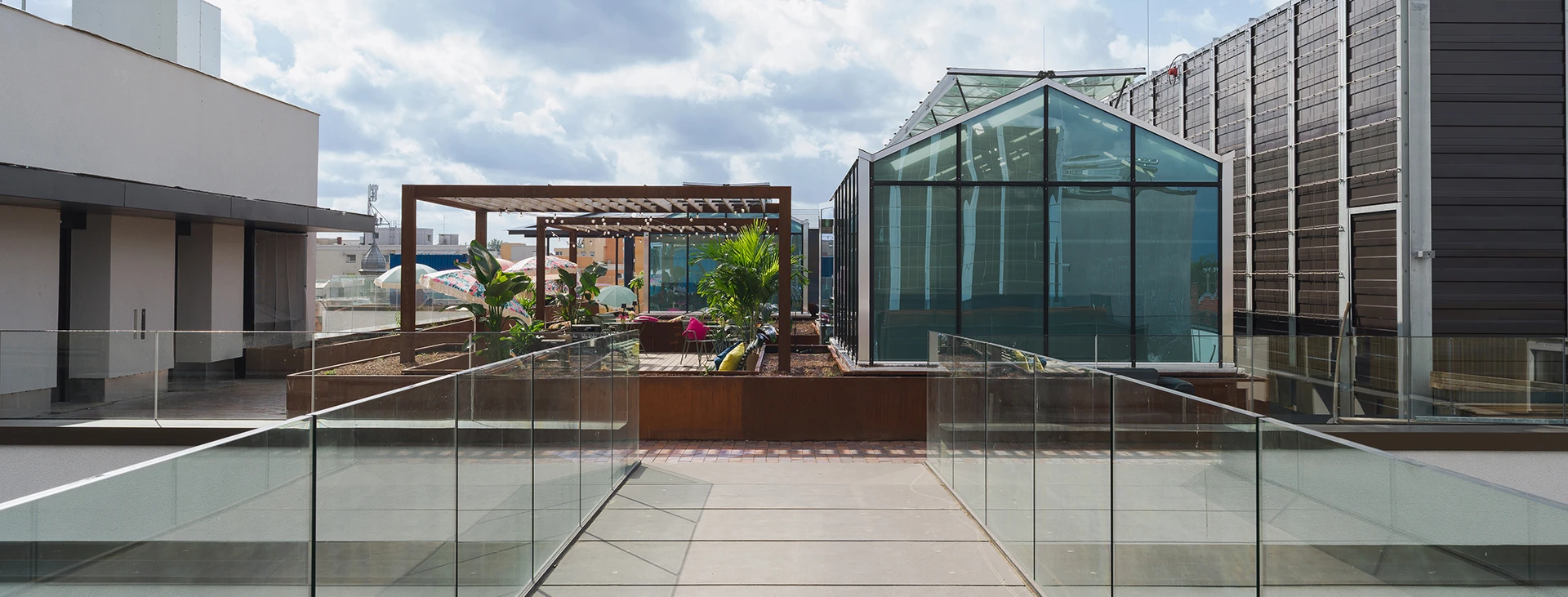 Moderne Dachterrasse mit Glasgebäude, umgeben von anderen Strukturen und Pflanzen unter einem bewölkten Himmel