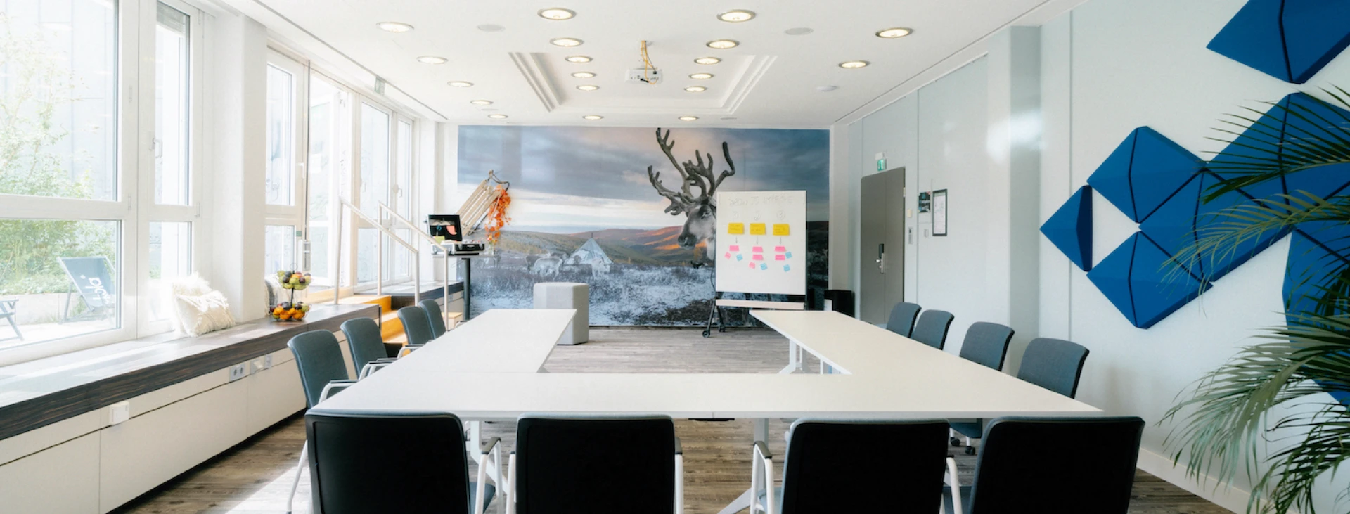 Moderner Konferenzraum mit U-förmiger Tischanordnung, großen Fenstern, Wandkunst mit Rentiermotiv und blauen Akustikpaneelen.