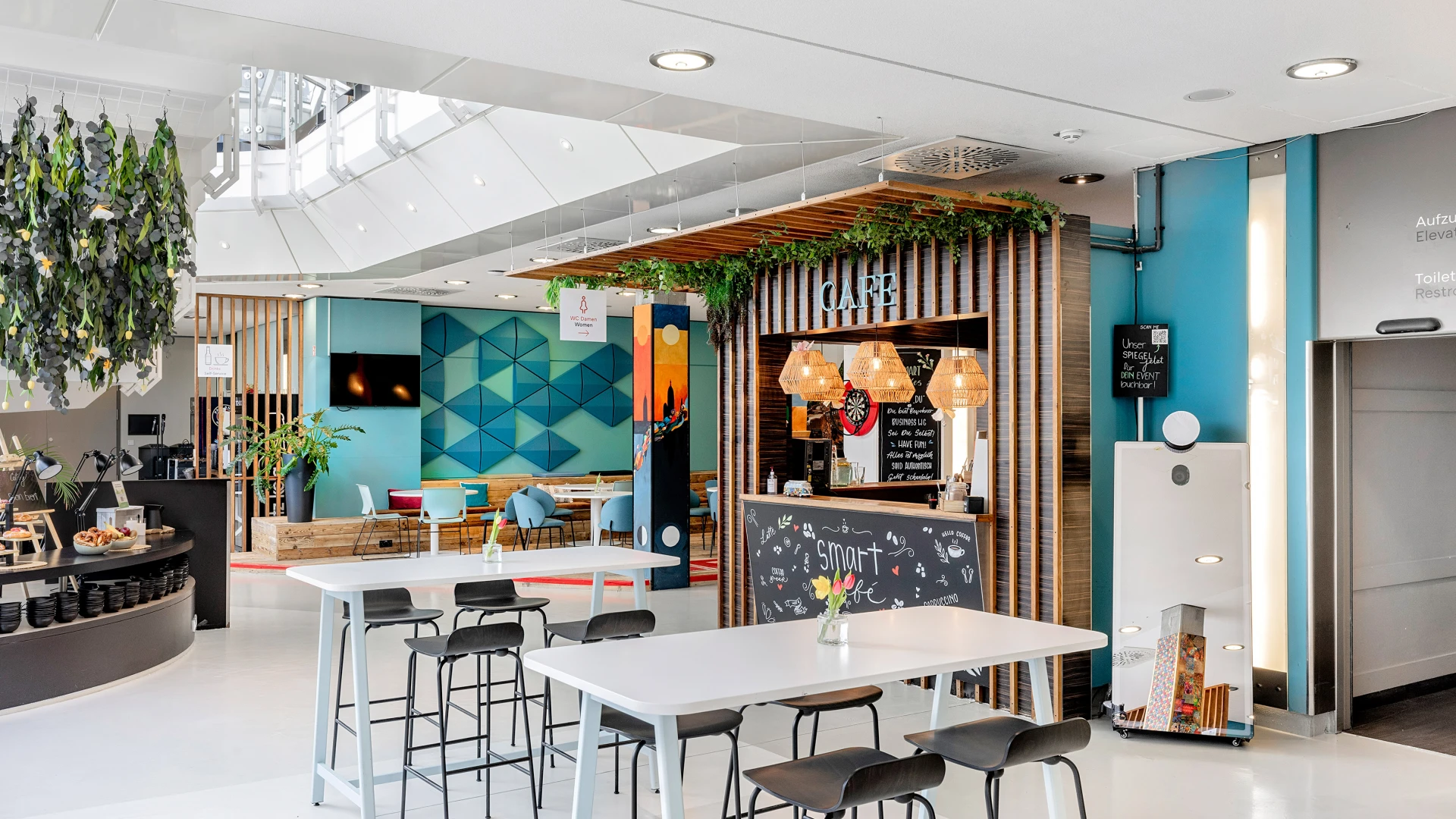 Bright, modern café with white tables and black bar stools, colorful wall décor, and greenery integrated into wooden elements. The café counter and seating area are visible in the background.