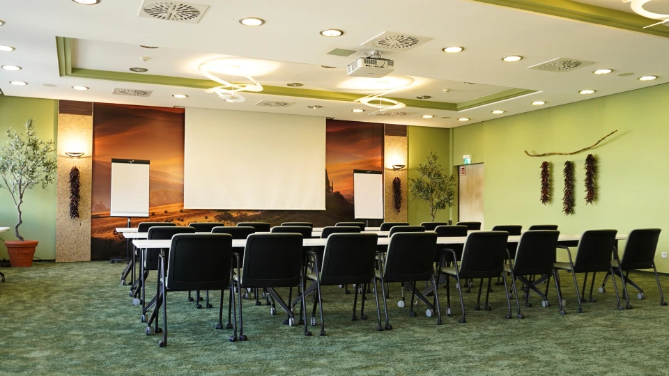 Leerer Seminarraum mit vielen schwarzen Stühlen um mehrere Tische angeordnet, großem Projektorbildschirm, dekorativen Pflanzen und Wandbildern.