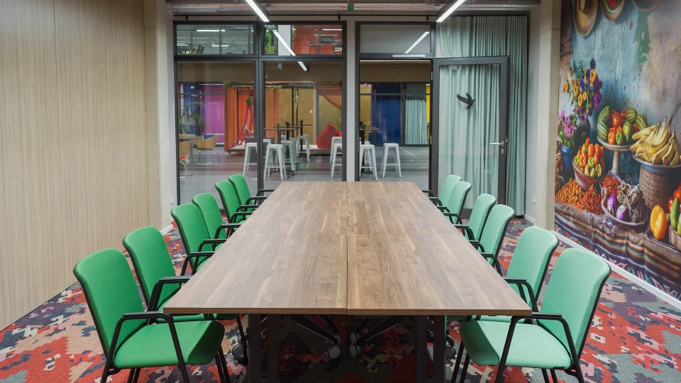 Innenansicht des Konferenzraums Kolumbien Bogota mit großem Holztisch, grünen Stühlen und Wandbild eines Obst- und Gemüsemarkts im Smartvillage München