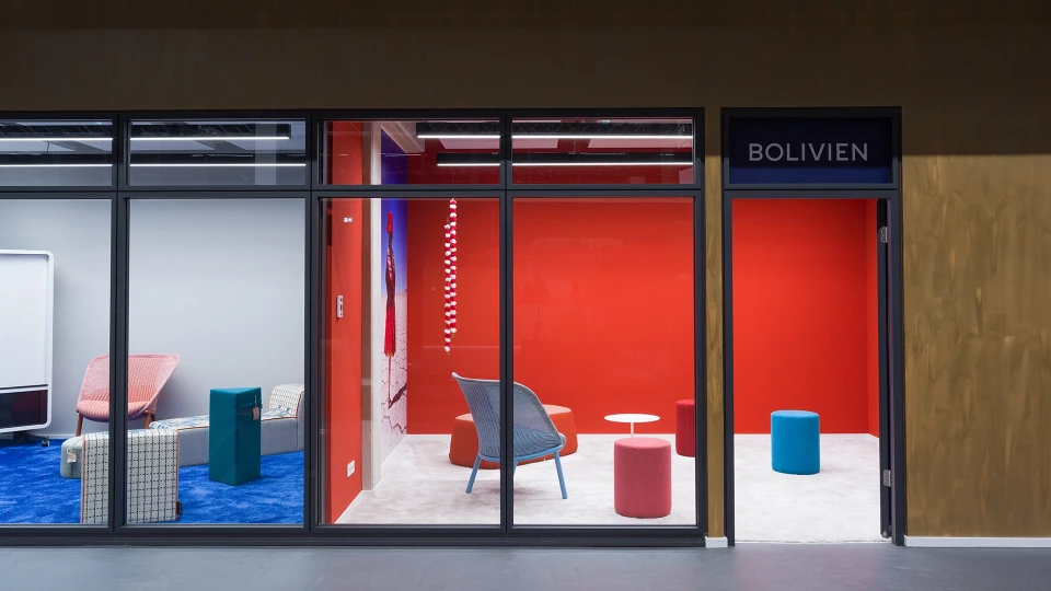 Moderner Lounge-Bereich im Raum Bolivien mit farbigen Sitzhockern, weißem Tisch und roter Wand im Smartvillage Köln