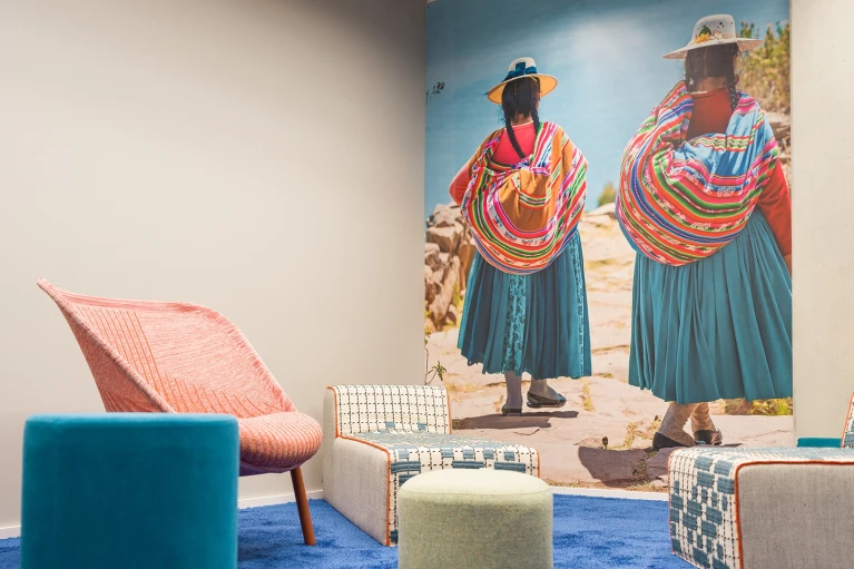 Cozy meeting room with colorful carpet, creative seating, and a wall painting of traditionally dressed women by a lake.