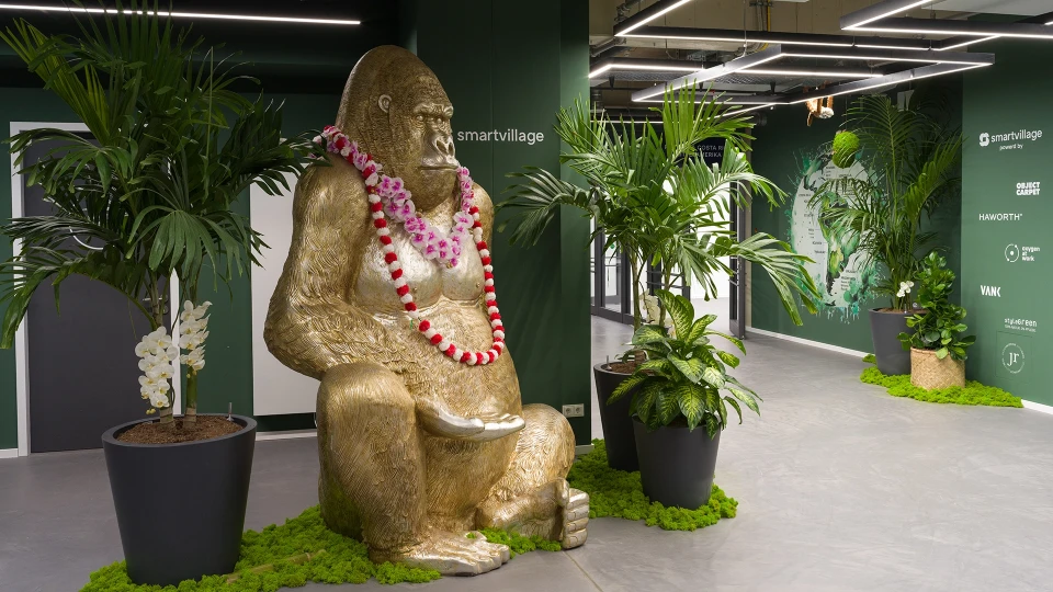Eine goldene Gorilla-Skulptur sitzt in einem Büroflur, geschmückt mit einer pinken Blumenkette, umgeben von mehreren großen Grünpflanzen.