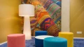 Moderne Sitzecke mit bunten runden Hockern in Pink, Gelb, Weiß und Blau auf einem dunkelblauen Teppich, daneben eine große weiße Stehlampe, vor einer Wand mit lebhaftem, mehrfarbigem Muster