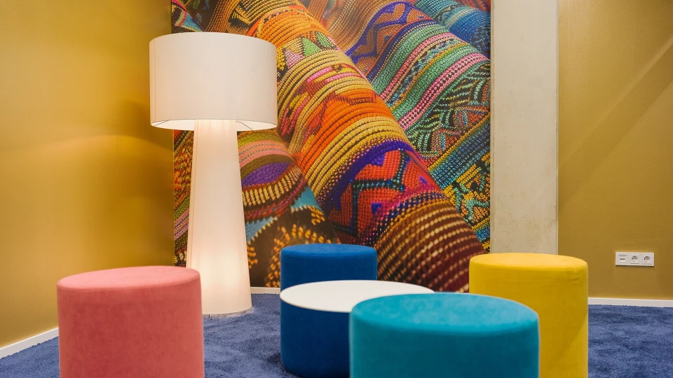 Moderne Sitzecke mit bunten runden Hockern in Pink, Gelb, Weiß und Blau auf einem dunkelblauen Teppich, daneben eine große weiße Stehlampe, vor einer Wand mit lebhaftem, mehrfarbigem Muster