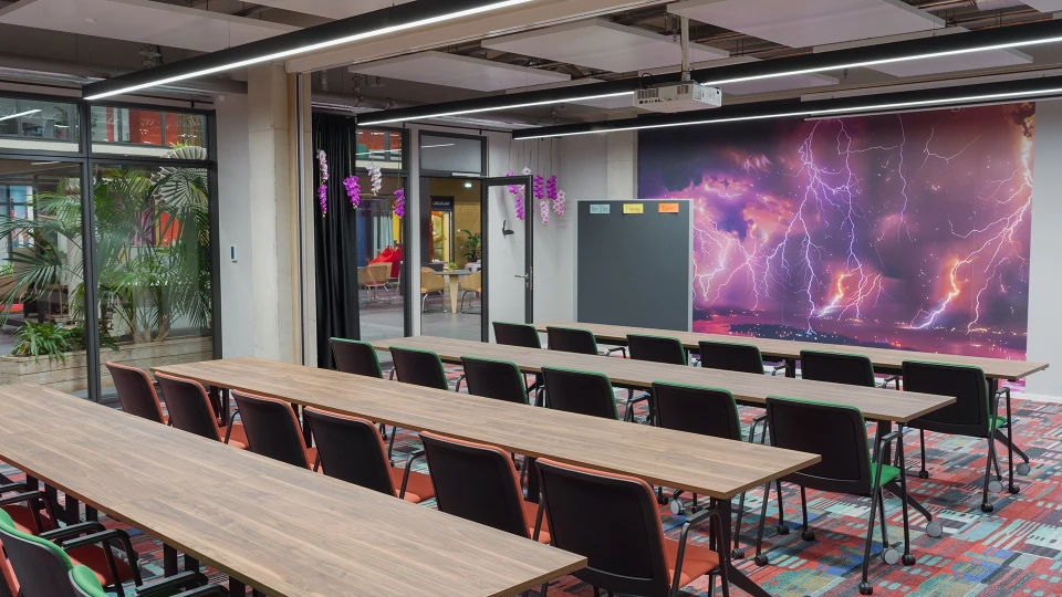 Modernes Bürokonferenzraum mit Holztischen und bunten Stühlen, dekoriert mit Zimmerpflanzen und einer großen Wandbildschirm mit Blitzdarstellung