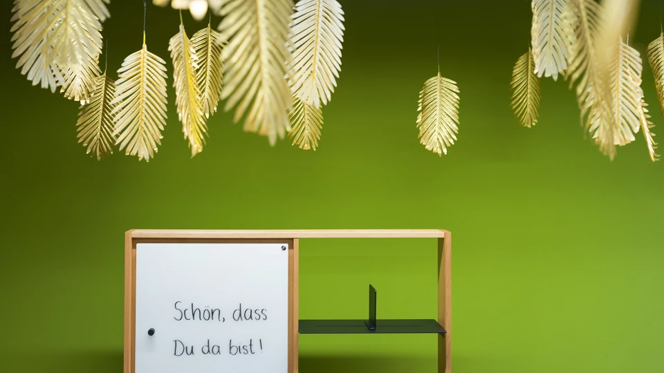 Blick durch das Fenster in den Raum "Guyana" mit grünem Wandanstrich, einer hellblauen Couch und weißer Stehlampe, umgeben von goldenen Blättern.