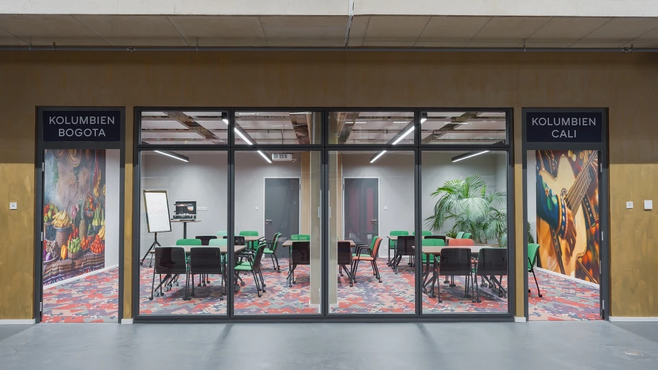 Seminarraum „Kolumbien Bogotá“ mit bunter Teppichstruktur, grünen und orangen Stühlen sowie einer großformatigen Wandgestaltung mit tropischen Früchten und Hüten.
