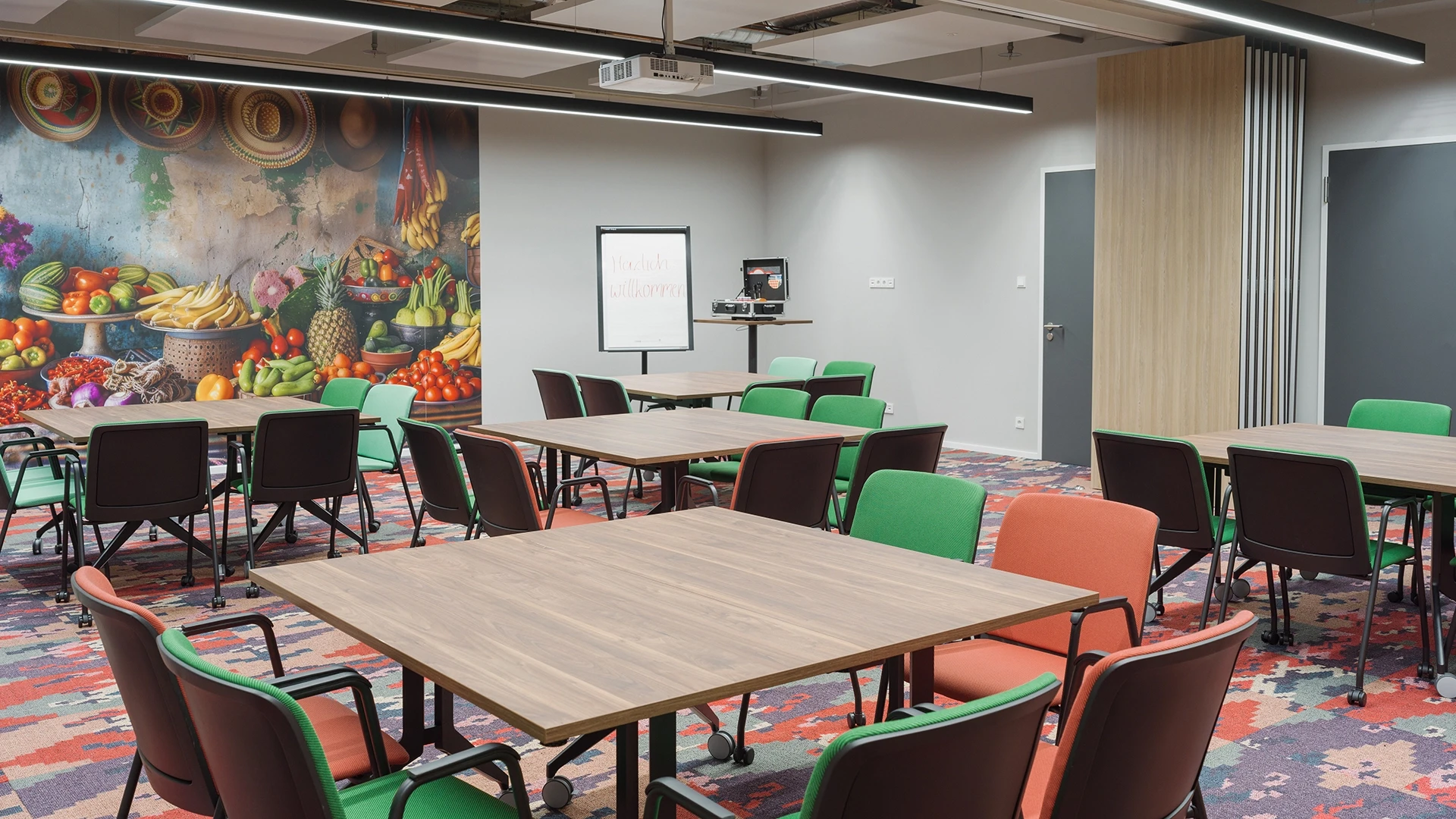 Seitlicher Blick auf den Raum „Kolumbien Bogotá“ mit Stuhlreihen vor der Obstmarkt-Wand, Flipchart mit Begrüßungstext und tragbarem Präsentationstool.
