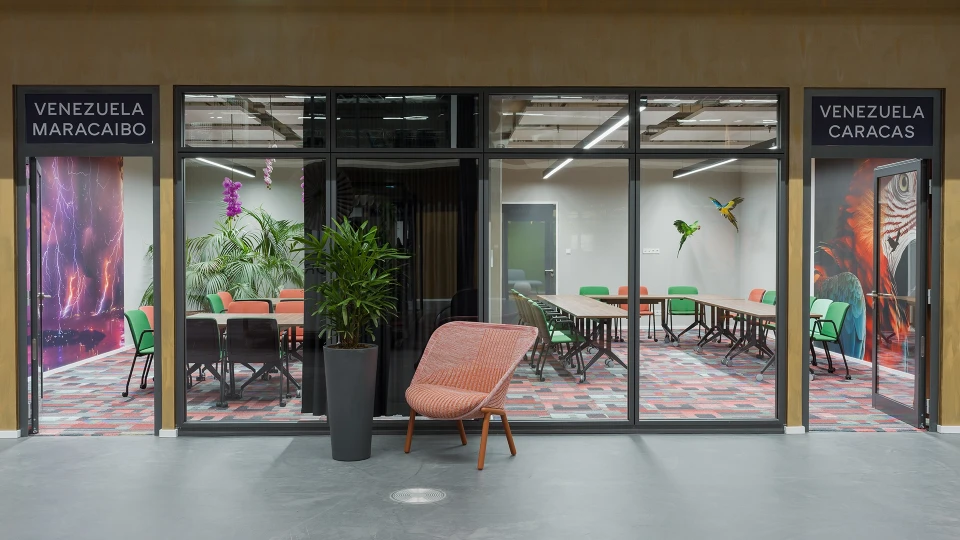 Moderne Büroumgebung hinter Glasfronten mit Beschriftung 'Venezuela Maracaibo' und 'Venezuela Caracas'. Innenraum mit bunten Stühlen, Tischen und Pflanzen.