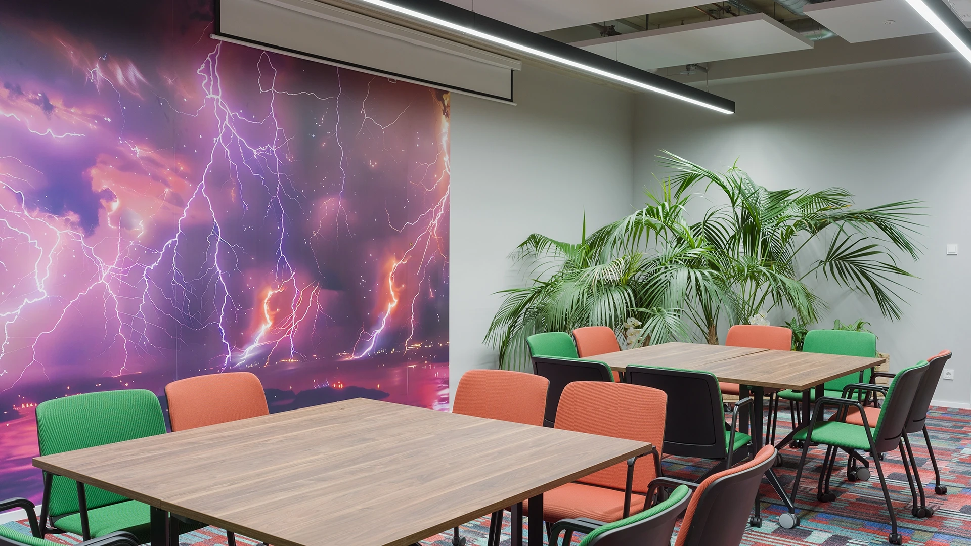 Besprechungsraum mit mehreren Tischen und Stühlen in Grün und Orange. Eine Wand ist mit einem großen Wandbild eines dramatischen lila Himmels mit Blitzen verziert.