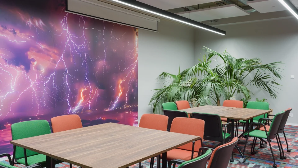 Besprechungsraum mit mehreren Tischen und Stühlen in Grün und Orange. Eine Wand ist mit einem großen Wandbild eines dramatischen lila Himmels mit Blitzen verziert.