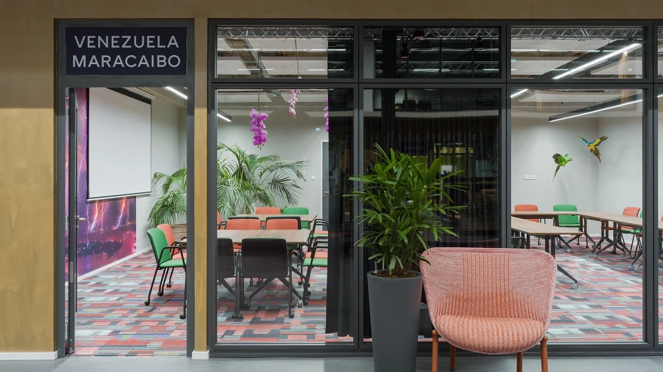 Modernes Bürointerieur mit großen Fenstern, Pflanzen und bunten Möbeln. Sicht auf einen Besprechungsraum mit Tischen, Stühlen und dekorativen Pflanzen.