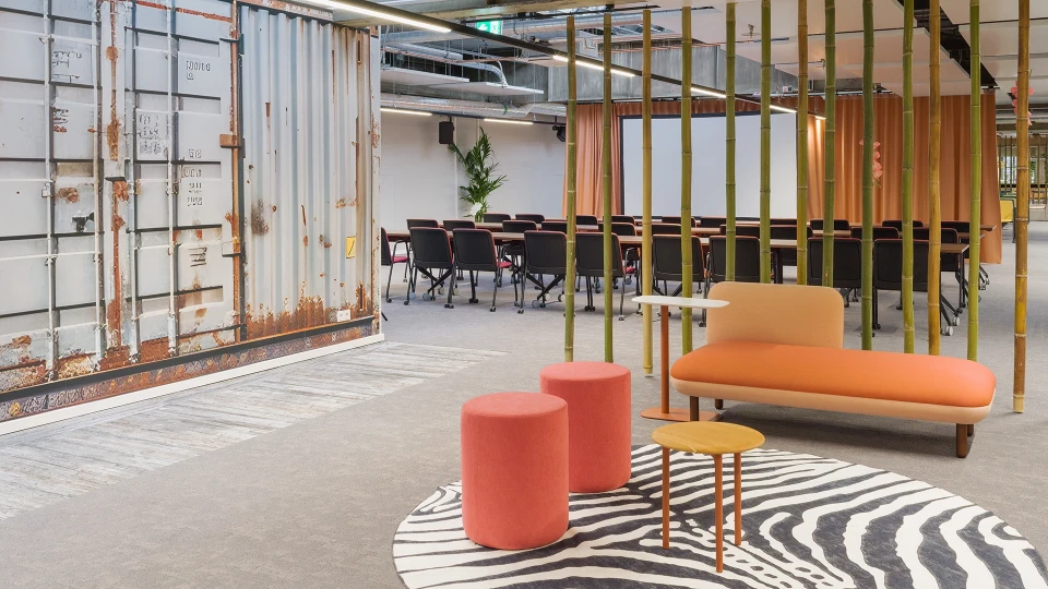 Lounge-Bereich mit orangener Bank, runden Hockern, Zebrateppich und Bambusstreben vor einem Konferenzbereich mit Kinobestuhlung.