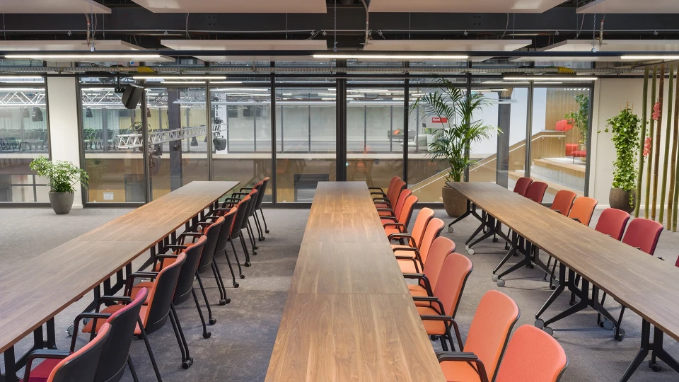 Konferenzbereich mit zwei langen Holztischen, umgeben von roten und orangenen Stühlen, große Fensterfront mit Blick auf Studio- oder Hallenbereich.