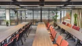Konferenzbereich mit zwei langen Holztischen, umgeben von roten und orangenen Stühlen, große Fensterfront mit Blick auf Studio- oder Hallenbereich.