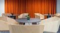Kleine Bistro-Ecke mit runden Tischen und modernen Holzschalenstühlen vor einem orangefarbenen Vorhang.