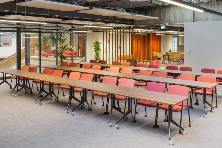 Seminar room with several rows of wheeled wooden tables and mixed color seating, with a bright lounge area in the background separated by bamboo partitions.