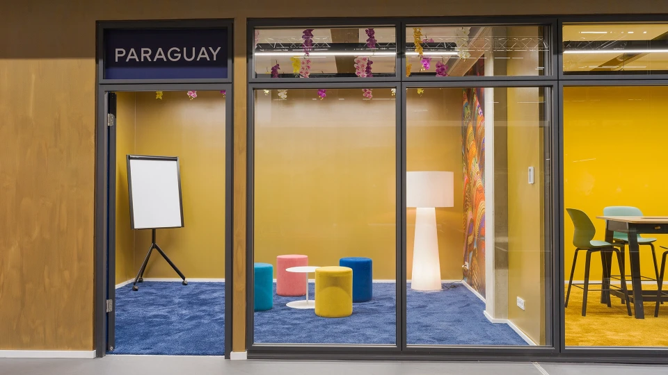 View through a glass wall into the 'Paraguay' room, featuring a colorful carpet, colored poufs, a white table, and a large floor lamp in front of a wall with a colorful textile pattern.