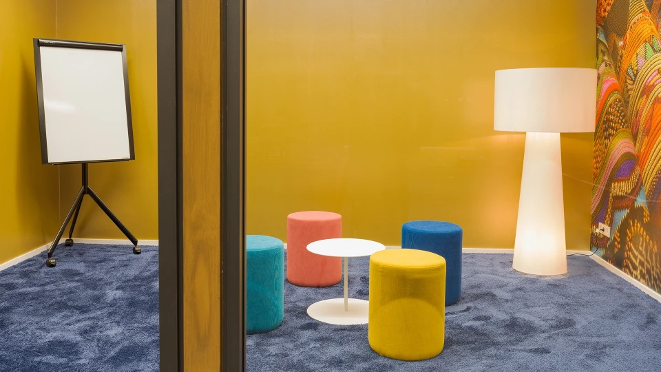 Close-up of five colorful poufs (yellow, turquoise, blue, orange, pink) around a round white side table on a dark carpet.