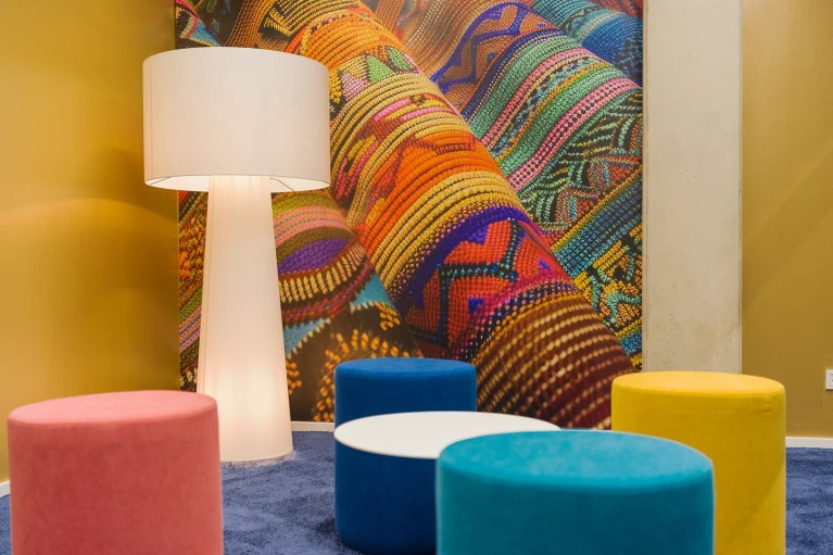 Interior view of the Paraguay Room with a tall floor lamp, colorful sitting stools, and a detailed wall graphic depicting vibrant textile fabric.