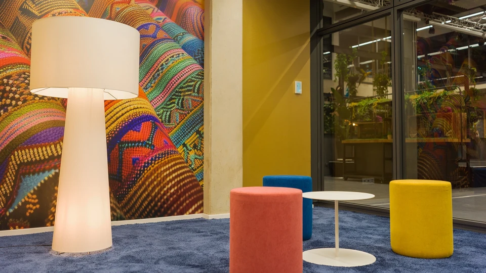 View through the window facade into the 'Paraguay' meeting room, featuring colorful wall design, blue carpet, and multicolored stools.