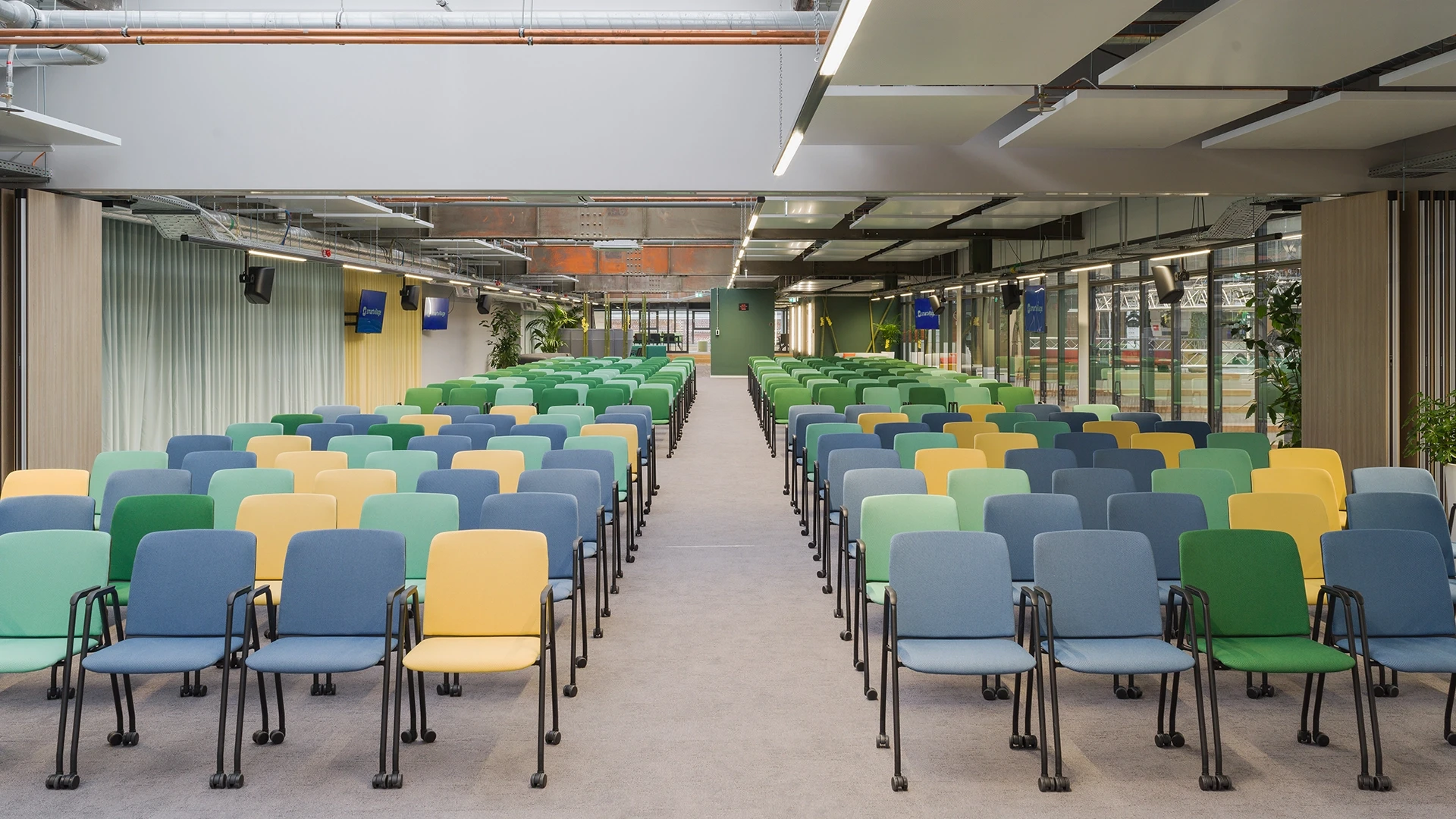 Central perspective view of a colorfully furnished event area with monitors and skylights.
